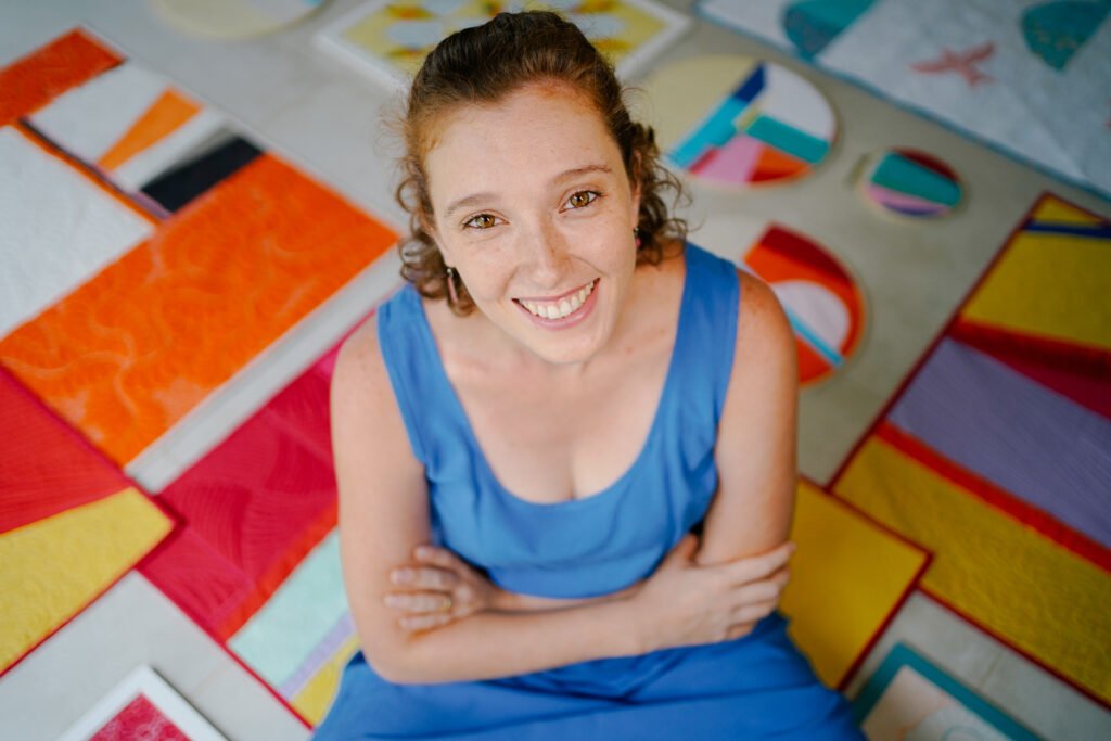 mulher de braços cruzados, sorrindo, com quilts modernos atrás dela, posicionados no chão